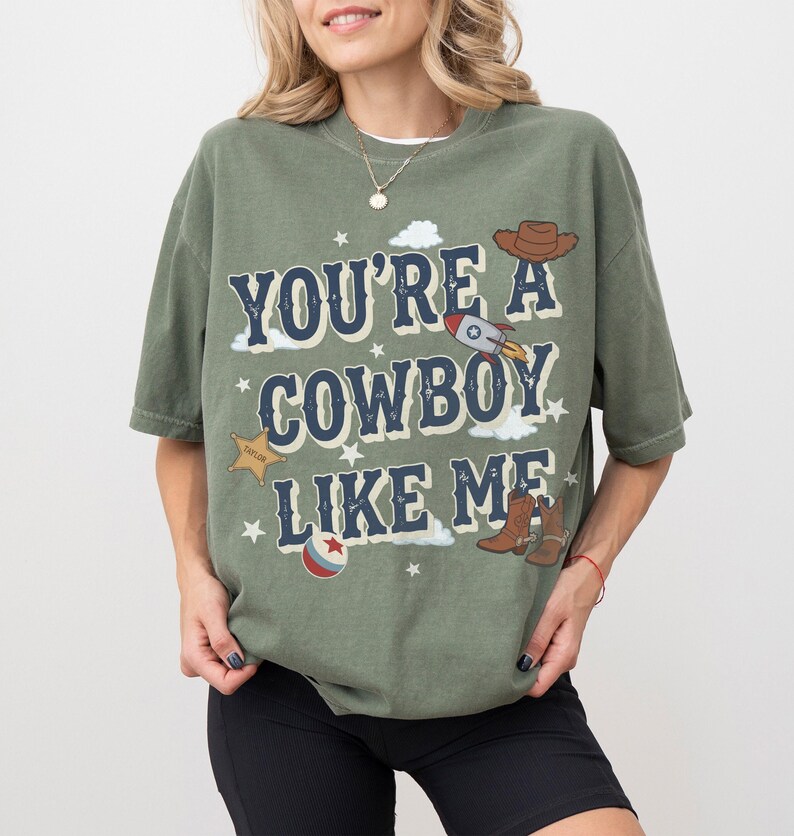 May include: An olive green short-sleeve t-shirt featuring the dark blue text "YOU'RE A COWBOY LIKE ME" with graphic illustrations of a brown cowboy hat, a rocket ship, a gold sheriff star, a pair of brown cowboy boots, and a red and blue ball.