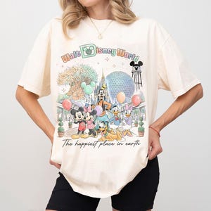 May include: Cream-colored t-shirt featuring a colorful graphic of Disney characters and the text "Walt Disney World." The design includes Mickey and Minnie Mouse, Goofy, Donald and Daisy Duck, and Pluto. The phrase "The happiest place in earth" is also printed on the shirt.