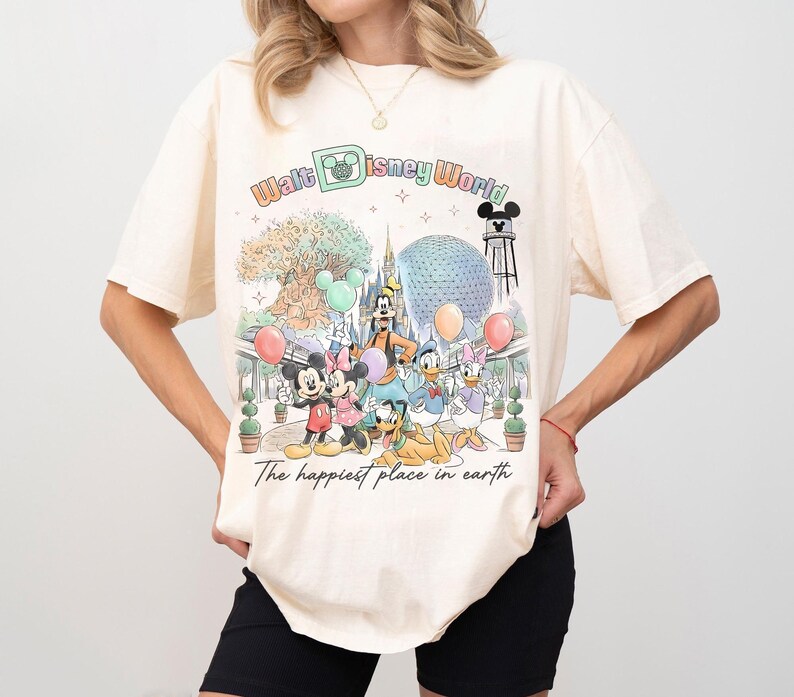 May include: Cream-colored t-shirt featuring a colorful graphic of Disney characters and the text "Walt Disney World." The design includes Mickey and Minnie Mouse, Goofy, Donald and Daisy Duck, and Pluto. The phrase "The happiest place in earth" is also printed on the shirt.