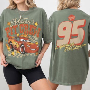 Op de afbeelding: Een olijfgroen t-shirt met een afbeelding van een rode raceauto en de tekst Mason's Pit Crew op de voorkant. De achterkant toont het nummer 95 in een groot beige lettertype met de tekst Lightning McQueen en een rood-wit Rust-eze logo.