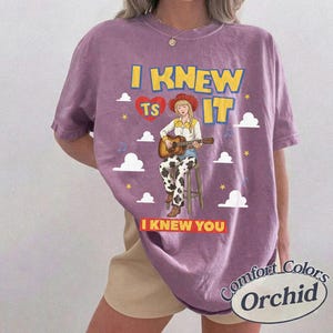 May include: A purple short-sleeve t-shirt featuring a graphic of a person in a cowboy hat playing an acoustic guitar while sitting on a stool. The text reads I KNEW TS IT and I KNEW YOU, surrounded by white clouds, yellow stars, and a red heart.
