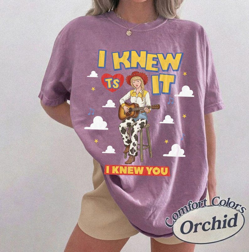 May include: A purple short-sleeve t-shirt featuring a graphic of a person in a cowboy hat playing an acoustic guitar while sitting on a stool. The text reads I KNEW TS IT and I KNEW YOU, surrounded by white clouds, yellow stars, and a red heart.