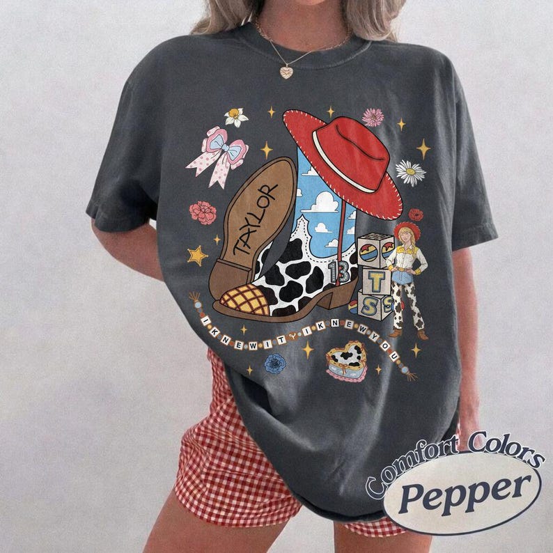 May include: A charcoal grey t-shirt featuring a graphic print of a cow-print cowboy boot with a red hat, a small character, blocks with the letters T, S, and the number 13, and the text TAYLOR and I KNEW IT I KNEW YOU in a circular arrangement.
