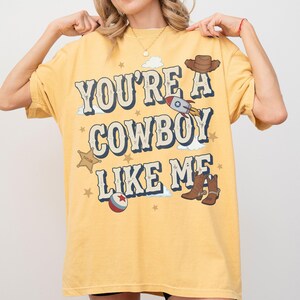 May include: A yellow short-sleeve t-shirt featuring the text "YOU'RE A COWBOY LIKE ME" in blue and white lettering. The graphic includes illustrations of a brown cowboy hat, a rocket ship, a sheriff badge, a ball, and brown cowboy boots.