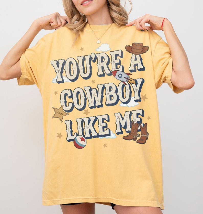 May include: A yellow short-sleeve t-shirt featuring the text "YOU'RE A COWBOY LIKE ME" in blue and white lettering. The graphic includes illustrations of a brown cowboy hat, a rocket ship, a sheriff badge, a ball, and brown cowboy boots.