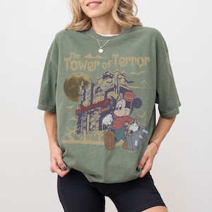 Puede incluir: Camiseta verde oliva con un gr&aacute;fico vintage de Mickey Mouse como botones, llevando una maleta, frente a un hotel embrujado. El texto "The Tower of Terror" est&aacute; impreso encima de la imagen. Una luna llena y p&aacute;jaros en el fondo.
