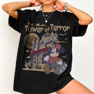 Puede incluir: Camiseta negra con un gr&aacute;fico de Mickey Mouse como botones, caminando frente a un hotel embrujado. El texto "The Tower of Terror" est&aacute; encima del hotel. Una luna llena y murci&eacute;lagos en el fondo.