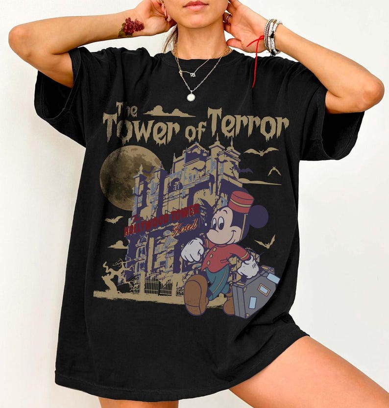 Puede incluir: Camiseta negra con un gr&aacute;fico de Mickey Mouse como botones, caminando frente a un hotel embrujado. El texto "The Tower of Terror" est&aacute; encima del hotel. Una luna llena y murci&eacute;lagos en el fondo.