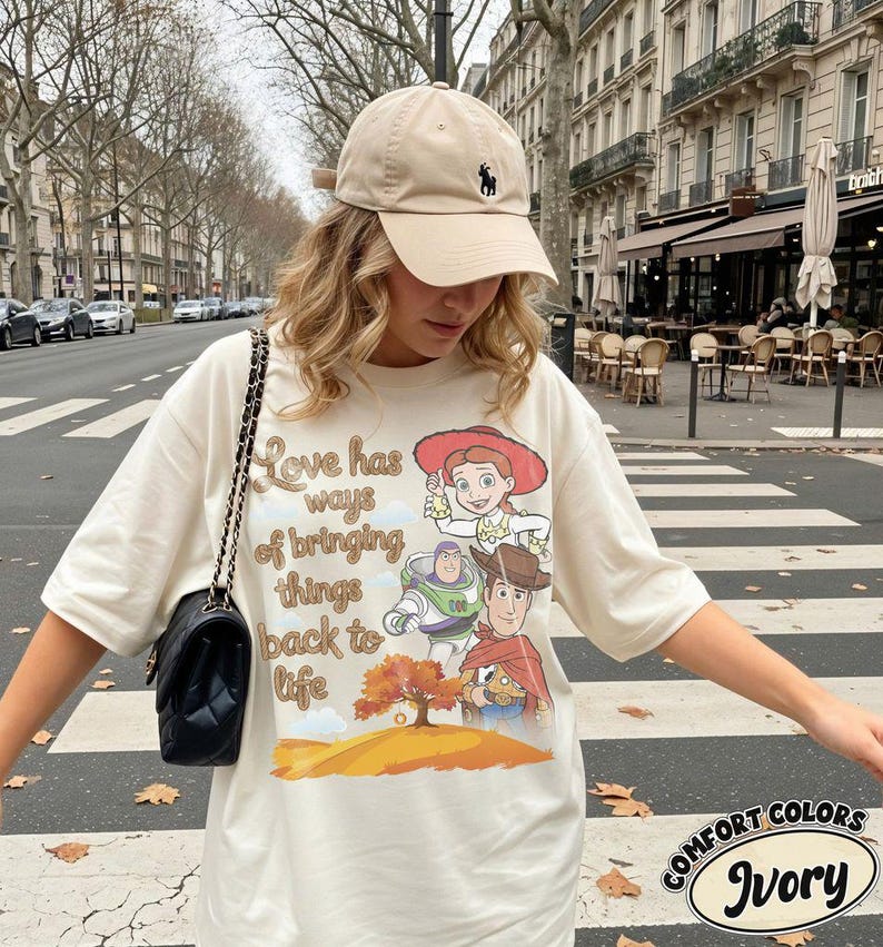 May include: An ivory t-shirt featuring a graphic print of Jessie, Buzz Lightyear, and Woody from Toy Story, with the text "Love has ways of bringing things back to life" above a tree illustration, paired with a beige baseball cap and a black bag.