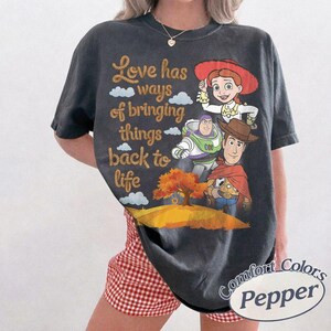 May include: A charcoal gray short-sleeve t-shirt featuring a graphic print of Jessie, Buzz Lightyear, and Woody from Toy Story. The text reads Love has ways of bringing things back to life above a landscape with a tree and a small swing.