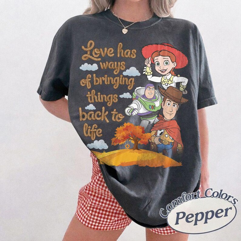May include: A charcoal gray short-sleeve t-shirt featuring a graphic print of Jessie, Buzz Lightyear, and Woody from Toy Story. The text reads Love has ways of bringing things back to life above a landscape with a tree and a small swing.