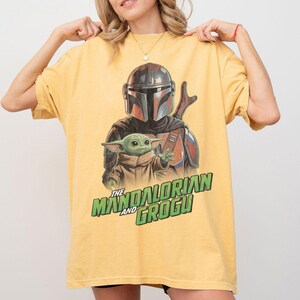May include: A yellow short-sleeve t-shirt featuring a graphic print of The Mandalorian and Grogu. The design shows the armored character standing behind the small green creature. The text THE MANDALORIAN AND GROGU is printed in green below.