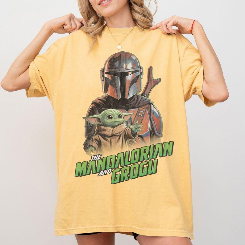 May include: A yellow short-sleeve t-shirt featuring a graphic print of The Mandalorian and Grogu. The design shows the armored character standing behind the small green creature. The text THE MANDALORIAN AND GROGU is printed in green below.