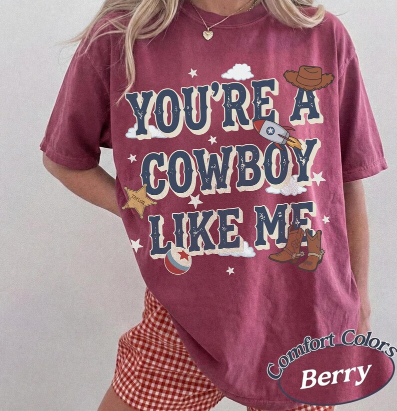 May include: A berry-colored short-sleeve t-shirt featuring the text "YOU'RE A COWBOY LIKE ME" in dark blue letters. The design includes graphics of a brown cowboy hat, a rocket ship, a gold sheriff star, a ball, and brown cowboy boots with stars.