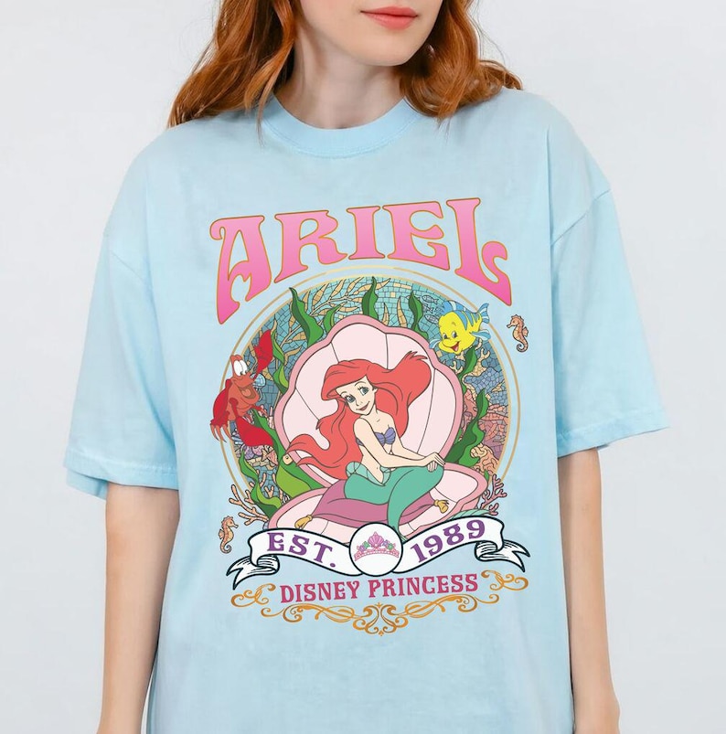 May include: Pale blue t-shirt with a graphic of Ariel from The Little Mermaid, seated in a seashell. The design includes the word "ARIEL" in pink, the year "1989", and the words "Disney Princess".