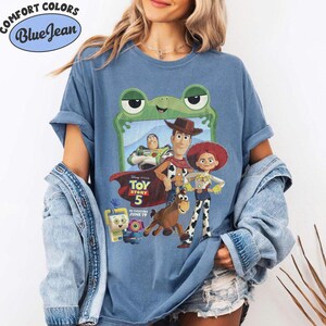 May include: A blue jean colored t-shirt featuring a graphic print of Toy Story 5 characters including Woody, Buzz Lightyear, Jessie, and Bullseye framed by a green frog outline. The text reads Disney Pixar Toy Story 5 In Theaters June 19.