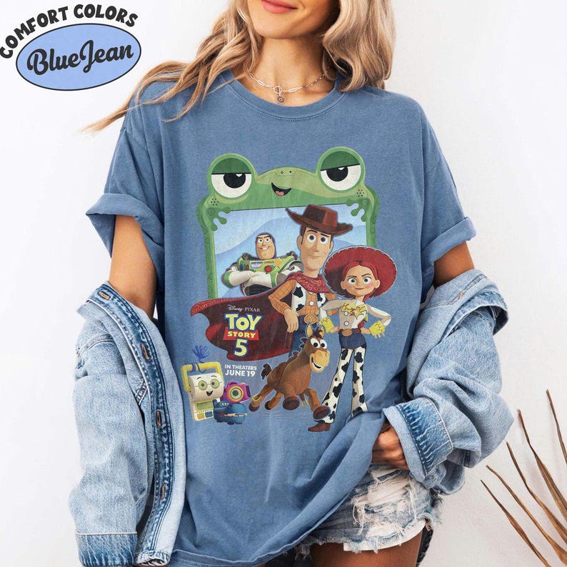 May include: A blue jean colored t-shirt featuring a graphic print of Toy Story 5 characters including Woody, Buzz Lightyear, Jessie, and Bullseye framed by a green frog outline. The text reads Disney Pixar Toy Story 5 In Theaters June 19.