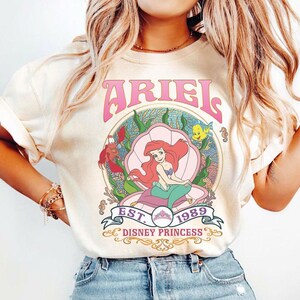 May include: Cream-coloured t-shirt with a graphic of Ariel from The Little Mermaid. The design includes the word "ARIEL" in pink, a seashell, and the text "EST. 1989 DISNEY PRINCESS". The illustration shows Ariel with red hair and a green tail.