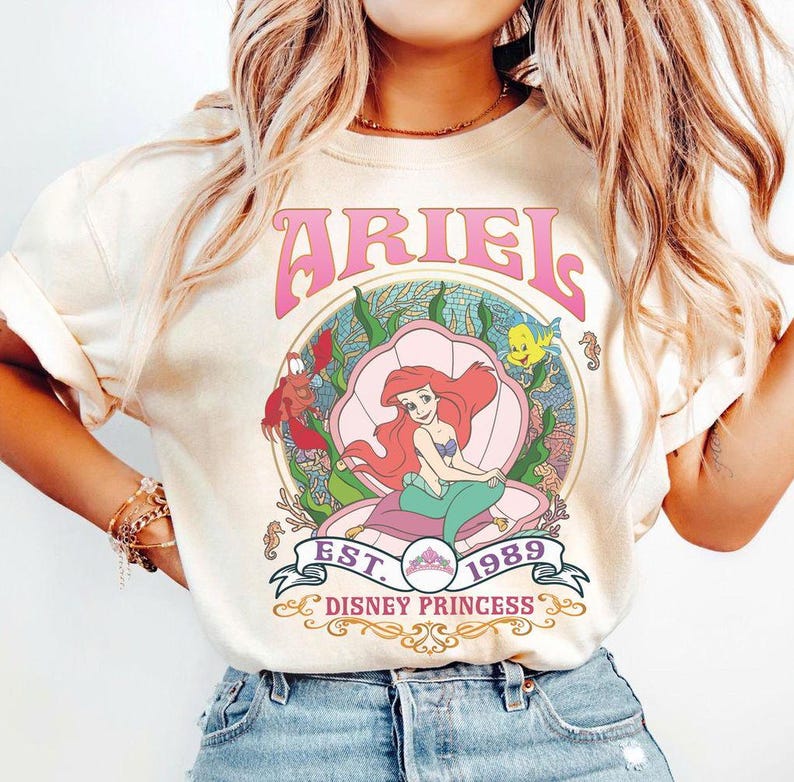 May include: Cream-coloured t-shirt with a graphic of Ariel from The Little Mermaid. The design includes the word "ARIEL" in pink, a seashell, and the text "EST. 1989 DISNEY PRINCESS". The illustration shows Ariel with red hair and a green tail.