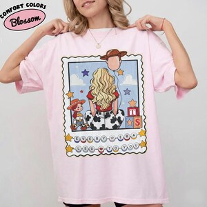 May include: A light pink t-shirt featuring a graphic print of a person with blonde hair, a cow-print skirt, and a cowboy hat. The design includes a small doll, building blocks, stars, clouds, and the text "EVERY ERA LED TO YOU" in circular beads.