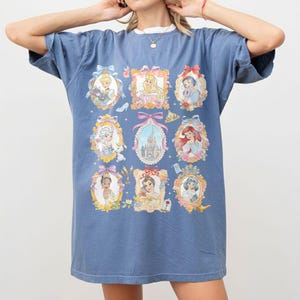 May include: A blue t-shirt featuring framed portraits of animated princesses. The portraits are in ornate gold frames with bows and floral accents. The shirt has a white collar and short sleeves.