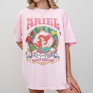 May include: Pale pink t-shirt with a graphic of Ariel from Disney's The Little Mermaid. The design includes the word "ARIEL" in pink, the phrase "EST. 1989 DISNEY PRINCESS", and an illustration of Ariel in a seashell.