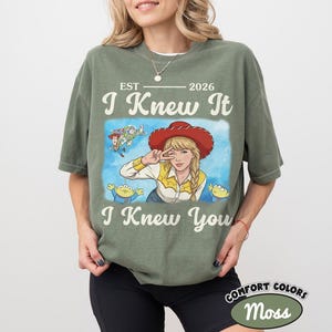 May include: A moss green short-sleeve t-shirt featuring a graphic print of a blonde character in a red cowboy hat, surrounded by small animated figures. The text reads EST 2026, I Knew It, and I Knew You in a cream-colored vintage-style font.
