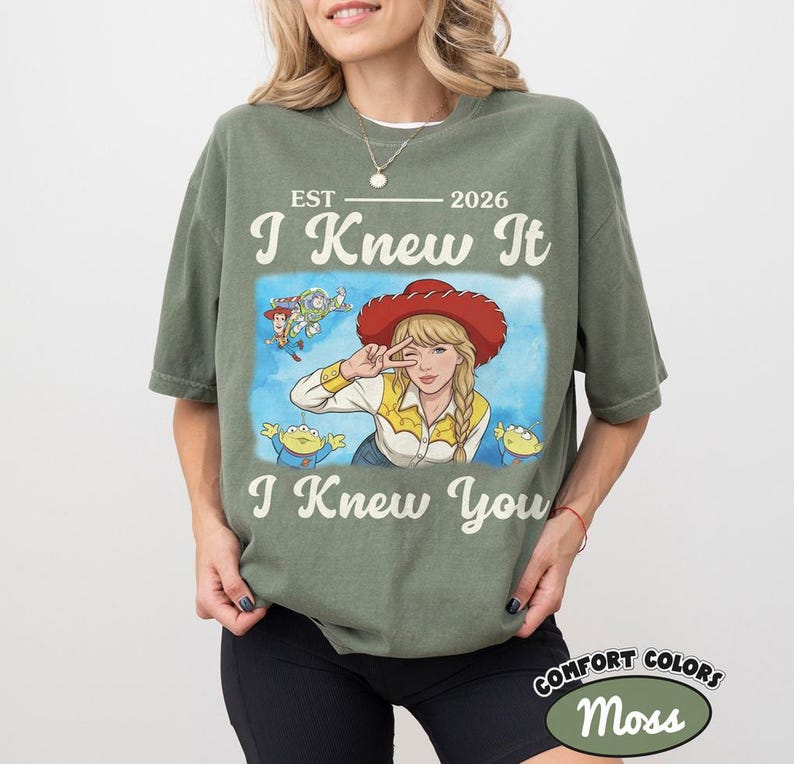 May include: A moss green short-sleeve t-shirt featuring a graphic print of a blonde character in a red cowboy hat, surrounded by small animated figures. The text reads EST 2026, I Knew It, and I Knew You in a cream-colored vintage-style font.