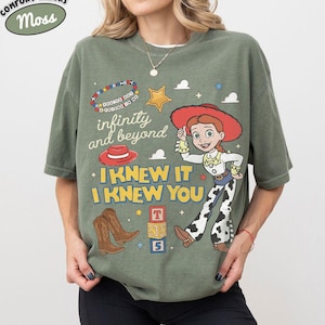 May include: An olive green t-shirt featuring a graphic print of a cowgirl character, brown cowboy boots, a red hat, a star, and alphabet blocks. The text reads "infinity and beyond" and "I KNEW IT I KNEW YOU" along with a beaded bracelet graphic.