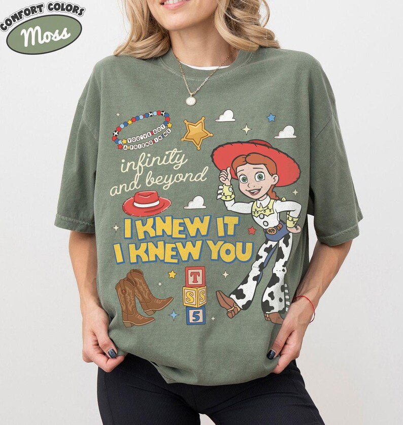 May include: An olive green t-shirt featuring a graphic print of a cowgirl character, brown cowboy boots, a red hat, a star, and alphabet blocks. The text reads "infinity and beyond" and "I KNEW IT I KNEW YOU" along with a beaded bracelet graphic.