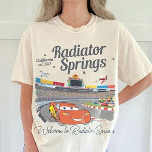 Puede incluir: Camiseta color crema con un gr&aacute;fico de Rayo McQueen corriendo en una pista en Radiator Springs. El dise&ntilde;o incluye el texto "Radiator Springs" y "California est. 2012". La camiseta tiene mangas cortas y cuello redondo.