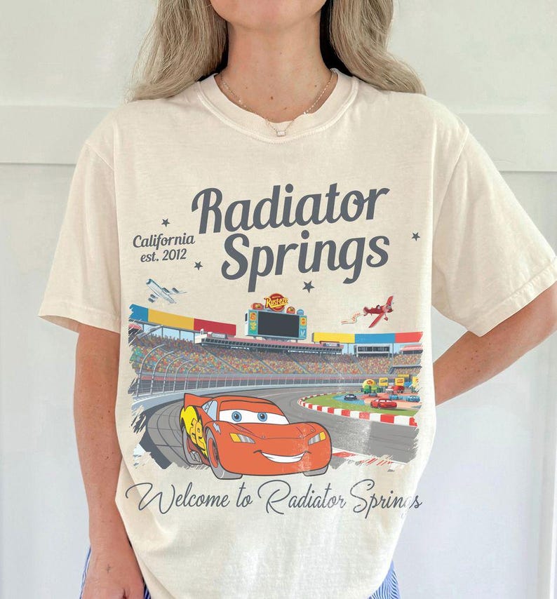 Puede incluir: Camiseta color crema con un gr&aacute;fico de Rayo McQueen corriendo en una pista en Radiator Springs. El dise&ntilde;o incluye el texto "Radiator Springs" y "California est. 2012". La camiseta tiene mangas cortas y cuello redondo.