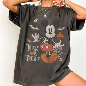 May include: A charcoal gray short-sleeve t-shirt featuring a graphic of a skeleton Mickey Mouse with red shorts, surrounded by Halloween elements including bats, a spider web, a pumpkin, candy, and the text Trick or Treat in white.