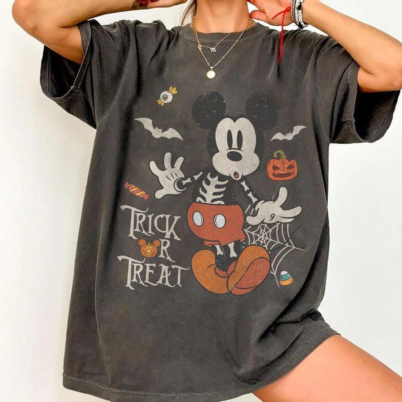 May include: A charcoal gray short-sleeve t-shirt featuring a graphic of a skeleton Mickey Mouse with red shorts, surrounded by Halloween elements including bats, a spider web, a pumpkin, candy, and the text Trick or Treat in white.