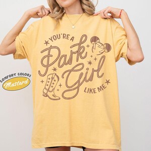 Op de afbeelding: Een mosterdgeel T-shirt met korte mouwen en bruine grafische tekst "YOU'RE A Park Girl LIKE ME", voorzien van illustraties van een cowboylaars en muizenoortjes. Het ontwerp bevat kleine steraccenten en letters in touwstijl.