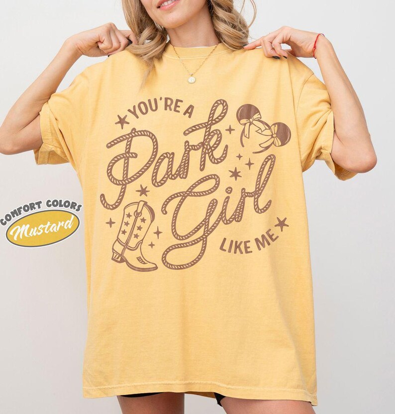 Op de afbeelding: Een mosterdgeel T-shirt met korte mouwen en bruine grafische tekst "YOU'RE A Park Girl LIKE ME", voorzien van illustraties van een cowboylaars en muizenoortjes. Het ontwerp bevat kleine steraccenten en letters in touwstijl.
