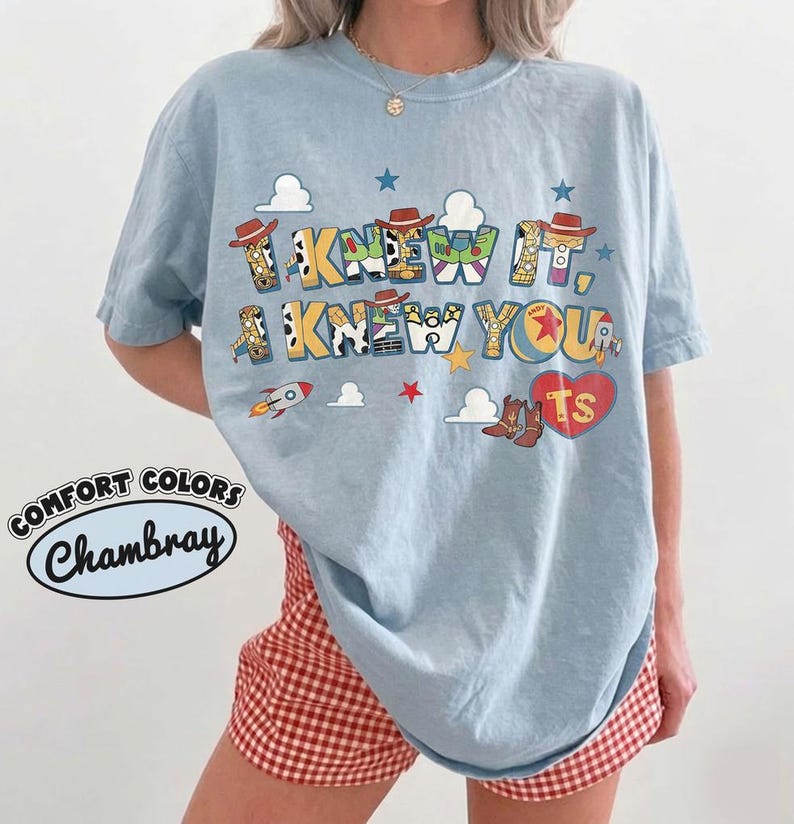 May include: A light blue short-sleeve t-shirt featuring a graphic print with the text "I KNEW IT, I KNEW YOU," decorated with cowboy hats, rockets, stars, clouds, and a red heart containing the letters "TS" in a colorful, patterned style.