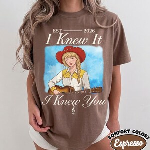 May include: A brown t-shirt featuring a graphic illustration of a person with blonde hair wearing a red cowboy hat and a yellow and white shirt while playing an acoustic guitar. The text reads EST 2026 I Knew It I Knew You with a treble clef.