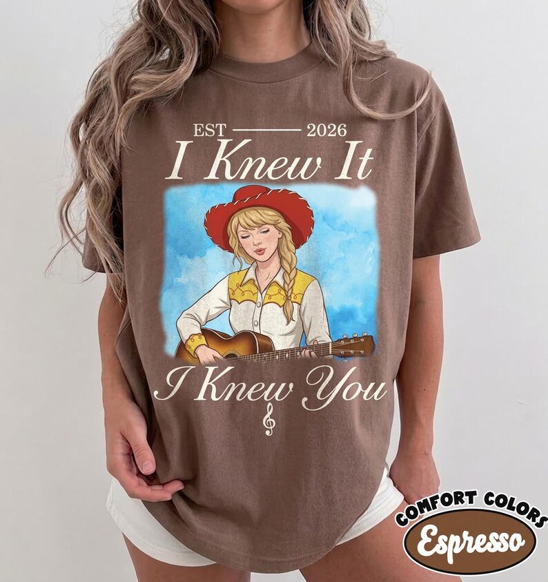 May include: A brown t-shirt featuring a graphic illustration of a person with blonde hair wearing a red cowboy hat and a yellow and white shirt while playing an acoustic guitar. The text reads EST 2026 I Knew It I Knew You with a treble clef.