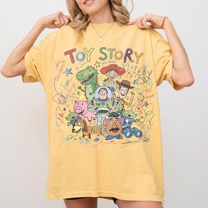 K&ouml;nnte beinhalten: Senfgelbes T-Shirt mit der Aufschrift "TOY STORY" und einer Grafik von Figuren aus dem Animationsfilm. Das Design umfasst Woody, Buzz Lightyear, Rex und andere Charaktere mit gekritzelten Akzenten.
