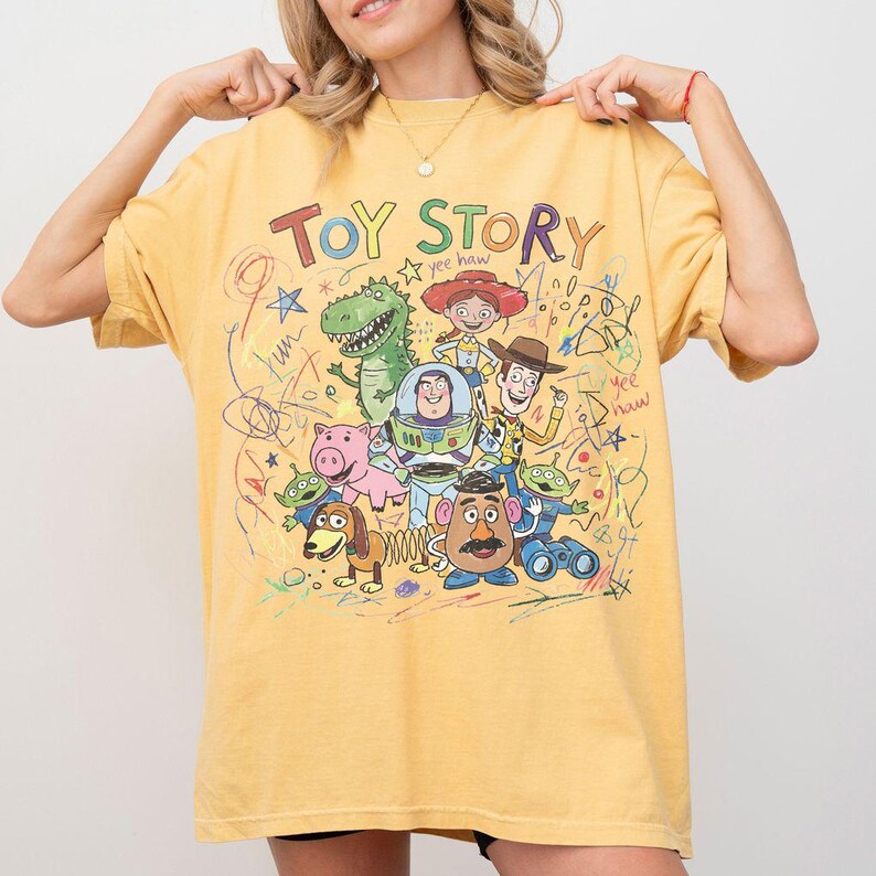 K&ouml;nnte beinhalten: Senfgelbes T-Shirt mit der Aufschrift "TOY STORY" und einer Grafik von Figuren aus dem Animationsfilm. Das Design umfasst Woody, Buzz Lightyear, Rex und andere Charaktere mit gekritzelten Akzenten.