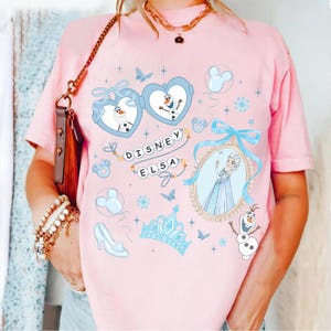 May include: A light pink t-shirt featuring Disney's Elsa and Olaf. The design includes heart-shaped frames with Olaf, a framed image of Elsa, and the text "DISNEY ELSA". Additional elements include butterflies, a crown, and a high heel.