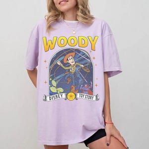 May include: A lilac t-shirt with the word "WOODY" in yellow above a graphic of Woody from Toy Story. The graphic includes the text "DISNEY" and "TOY STORY" on a banner.
