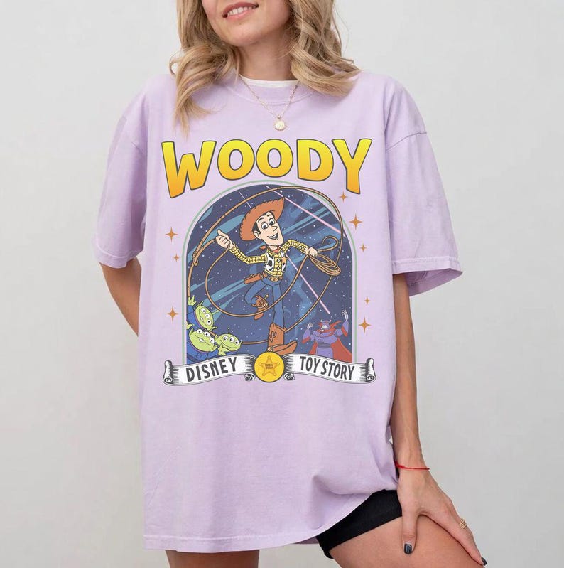 May include: A lilac t-shirt with the word "WOODY" in yellow above a graphic of Woody from Toy Story. The graphic includes the text "DISNEY" and "TOY STORY" on a banner.