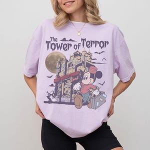 Puede incluir: Camiseta lila claro con un dise&ntilde;o de Mickey Mouse frente a la Torre del Terror. El texto "The Tower of Terror" est&aacute; sobre la imagen. El dise&ntilde;o incluye una luna llena y murci&eacute;lagos.