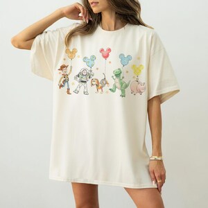 May include: Cream-colored oversized t-shirt featuring a colorful graphic of animated characters holding Mickey Mouse-shaped balloons. The design includes a cowboy, a space ranger, a dog, a dinosaur, and a pig. The shirt has short sleeves and a relaxed fit.