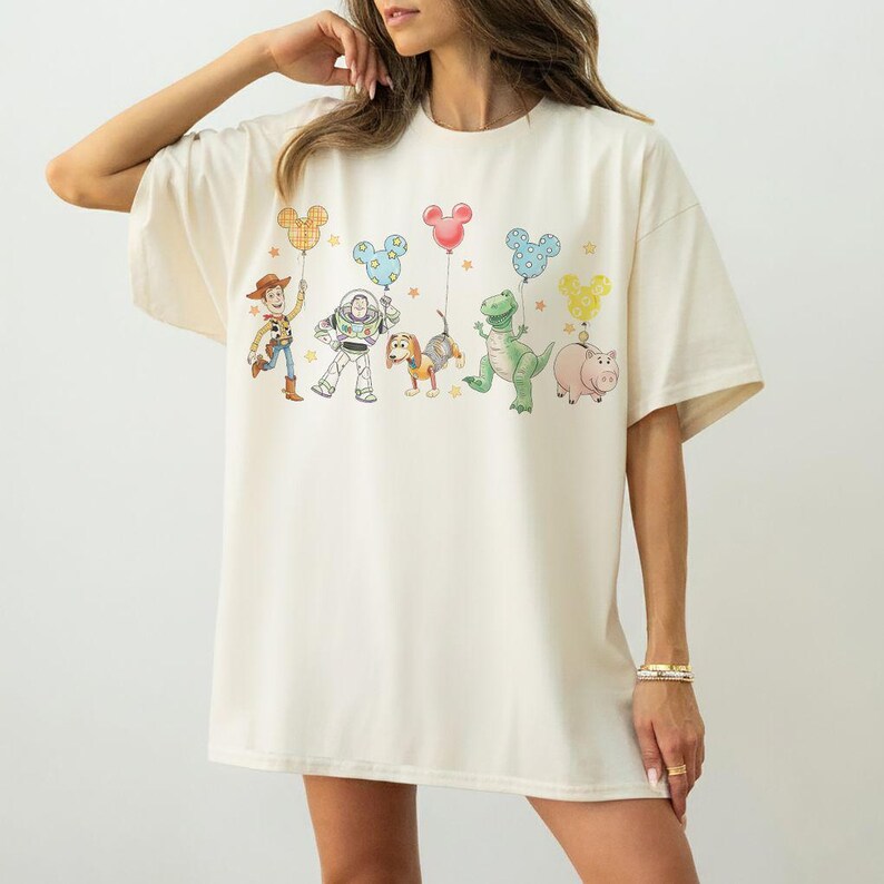 May include: Cream-colored oversized t-shirt featuring a colorful graphic of animated characters holding Mickey Mouse-shaped balloons. The design includes a cowboy, a space ranger, a dog, a dinosaur, and a pig. The shirt has short sleeves and a relaxed fit.