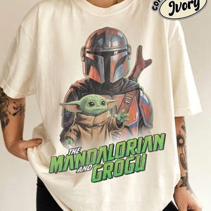 May include: An ivory t-shirt featuring a graphic print of The Mandalorian and Grogu. The design shows the armored character behind the small green creature. Below the characters, the text reads THE MANDALORIAN AND GROGU in green letters.