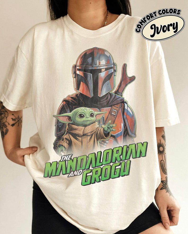 May include: An ivory t-shirt featuring a graphic print of The Mandalorian and Grogu. The design shows the armored character behind the small green creature. Below the characters, the text reads THE MANDALORIAN AND GROGU in green letters.
