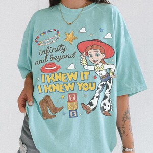 May include: A light blue t-shirt featuring a graphic print of the character Jessie from Toy Story, brown cowboy boots, a red hat, a yellow star, and colorful blocks. The text reads infinity and beyond, I KNEW IT, I KNEW YOU, and YOU'VE GOT A FRIEND IN ME.